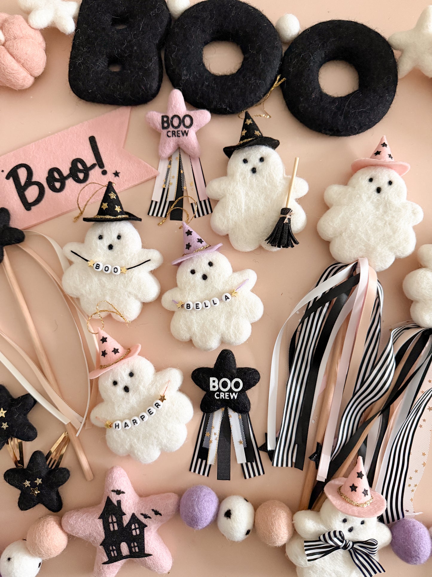 A flat-lay collection of handmade Halloween-themed felt ornaments and accessories, including ghost ornaments, witch hats, a star-shaped house, a 'BOO CREW' sign, and a 'Boo!' banner, arranged on a light pink background.