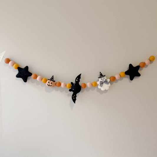 A Halloween-themed felt garland hanging on a plain light gray wall. The garland features orange, peach, and white felt balls, black star, a bat, cheeky  ghost and pumpkin details.