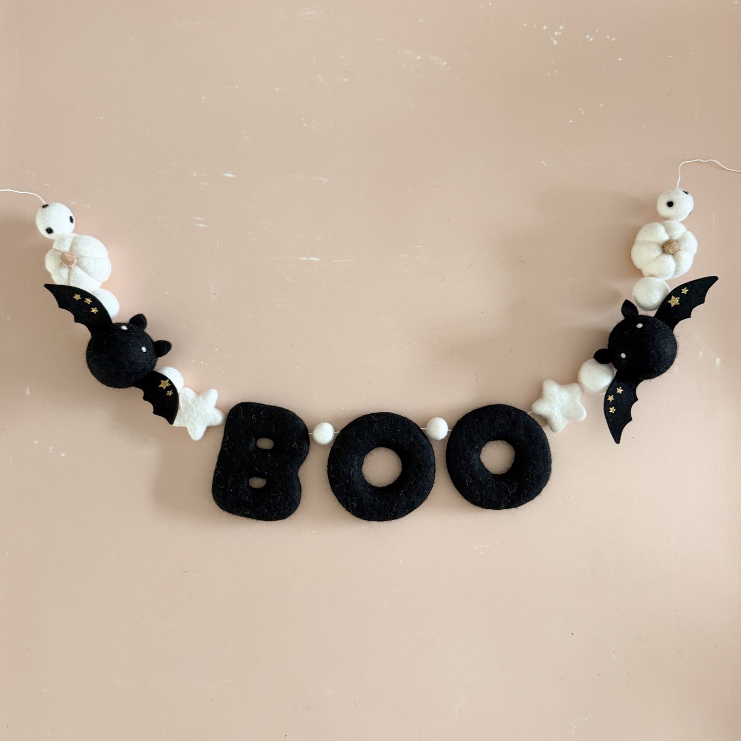 A Halloween-themed felt garland spelling out 'BOO' in black letters, decorated with black felt bats, white felt stars, and white pumpkins, hanging against a solid beige background.